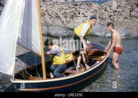 Cornwall, England. 1964 Uhr – drei junge Männer manövrieren ein kleines Segelboot auf den Strand. Zwei tragen Rettungswesten. Stockfoto