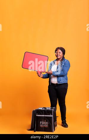 Porträt einer Zustellerin mit roter Sprechblase, die im Studio mit gelbem Hintergrund für Essen zum Mitnehmen wirbt. Restauran Kurier mit Thermal-Rucksack Stockfoto