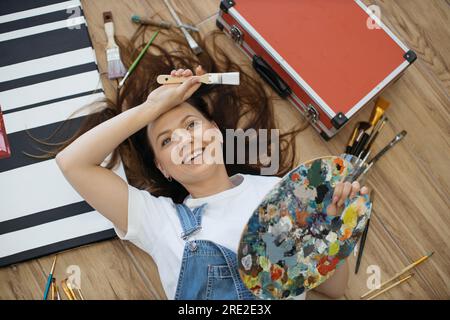 Junge, traumhafte Dame mit Palette und Pinsel, während sie in der Werkstatt unter Künstlerwerkzeugen lag. Talentierter Designer in Jeans und weißem T-Shirt, der neue Projekte im Sinn hat, während er sich auf dem Boden in der Halle entspannt. Stockfoto