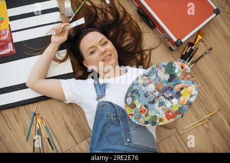 Junge, traumhafte Dame mit Palette und Pinsel, während sie in der Werkstatt unter Künstlerwerkzeugen lag. Talentierter Designer in Jeans und weißem T-Shirt, der neue Projekte im Sinn hat, während er sich auf dem Boden in der Halle entspannt. Stockfoto