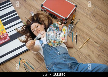 Junge, traumhafte Dame mit Palette und Pinsel, während sie in der Werkstatt unter Künstlerwerkzeugen lag. Talentierter Designer in Jeans und weißem T-Shirt, der neue Projekte im Sinn hat, während er sich auf dem Boden in der Halle entspannt. Stockfoto