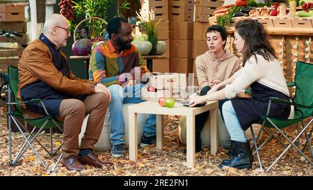 Glückliche Gruppe von Freunden, die Produktverkostungen auf dem lokalen Lebensmittelmarkt machen, treffen sich auf der Messe. Vielfältige Menschen genießen den Geschmack von Bio-saftigen Äpfeln, trinken hausgemachten Wein mit Bauern. Handgeführte Aufnahme. Stockfoto