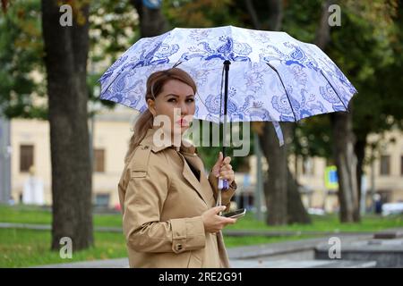 Asiatische Frau mit Schirm und Smartphone, die auf einer Straße steht. Regenwetter in der Stadt, Sommersturm Stockfoto