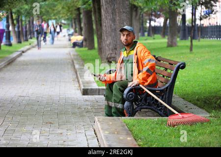 Hausmeister sitzt mit Smartphone auf einer Bank im Sommerpark. Arbeiter der öffentlichen Versorgungsunternehmen während der Ruhezeit, Straßenreinigung Stockfoto