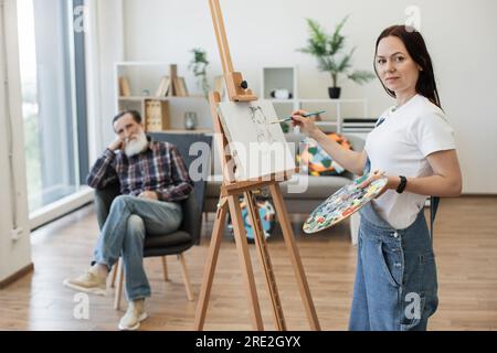 Selbstständige Maler-Maler in JeansOveralls mit Kopf-und-Schulter-Darstellung des Sitters in modernen Studioräumen. Attraktive brünette Dame, die einen Roug skizziert Stockfoto