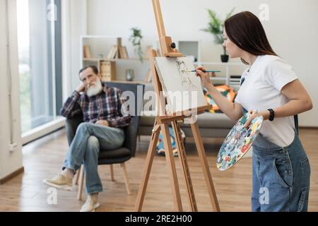 Selbstständige Maler-Maler in JeansOveralls mit Kopf-und-Schulter-Darstellung des Sitters in modernen Studioräumen. Attraktive brünette Dame, die einen Roug skizziert Stockfoto