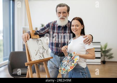 Fröhliche ältere Person, die eine glückliche weiße Dame nach erfolgreicher Kreativität im Studioinnenraum umarmt. Stolze Kunstlehrerin gratulierte der Studentin Stockfoto