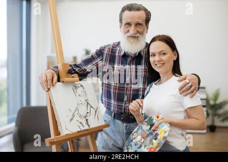 Fröhliche ältere Person, die eine glückliche weiße Dame nach erfolgreicher Kreativität im Studioinnenraum umarmt. Stolze Kunstlehrerin gratulierte der Studentin Stockfoto