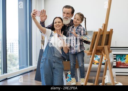 Eine reizende Familie von drei Generationen, die Selfie auf einem persönlichen Smartphone macht und Bilder auf einer hölzernen Staffelei malt. Mutter, Tochter und Großvater haben sich am Wochenende in einer geräumigen Wohnung amüsiert. Stockfoto