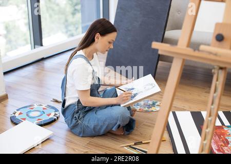 Seitenansicht einer fokussierten Malerin, die einen Jeansanzug trägt, auf einem Holzboden sitzt und im eigenen Kunststudio an einer neuen Zeichnung arbeitet. Junge Dame, die für Stockfoto