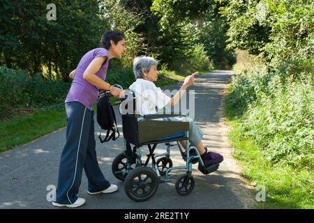 Asiatische Indianerin schubst ihre ältere Mutter im Rollstuhl im Sommer in Großbritannien nach draußen. Stellen Sie möglicherweise auch einen Betreuer, Fürsorge in der Gemeinde dar Stockfoto