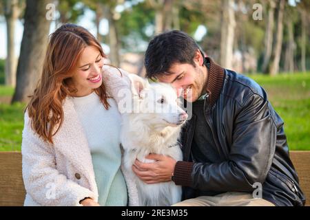 Ein junges weißes Paar streichelt seinen Hund auf einer Bank in einem Park. Stockfoto