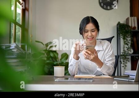 Eine attraktive asiatische Geschäftsfrau, die mit ihren Freunden Textnachrichten schreibt oder E-Mails auf ihrem Smartphone abruft, während sie an ihrem Schreibtisch im Büro sitzt. Stockfoto