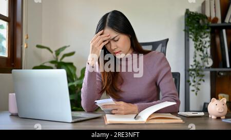 Junge Asiatin, die sich aufgebracht und gestresst fühlt, nachdem sie sich die Rechnungen angesehen hat. Papierdokumente verwalten, Rechnungen oder Steuern bezahlen. Stockfoto