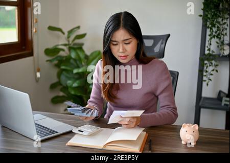 Eine junge asiatische Frau, die ihre Steuern und monatlichen Rechnungen berechnet, den Taschenrechner auf ihrem Schreibtisch verwendet und versucht, ihr Budget für ihre Einsparungen zu sparen Stockfoto