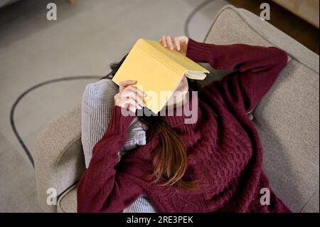 Junge Asiatin, die sich auf dem Sofa mit dem Bucheinband im Gesicht hingelegt hat, ein Nickerchen macht und sich in der Freizeit entspannt Stockfoto