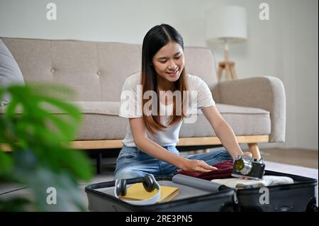 Eine glückliche und wunderschöne Asiatin bereitet ihre Sachen vor und packt ihr Gepäck in ihr Wohnzimmer, um sich auf ihre Auslandsreise vorzubereiten. Reisen, j Stockfoto