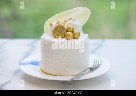 Kokosnusskuchen dekoriert mit weißer Schokolade, Keksen und Blattgold auf einem weißen Teller aus Granit mit Gartenhintergrund. Stockfoto