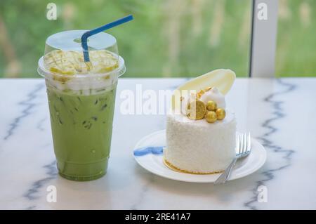 Grüner Eismatcha-Tee in Plastikbecher und Kokosnusskuchen dekoriert mit weißer Schokolade, Keksen und Blattgold auf einem weißen Teller aus Granit mit einem Stockfoto
