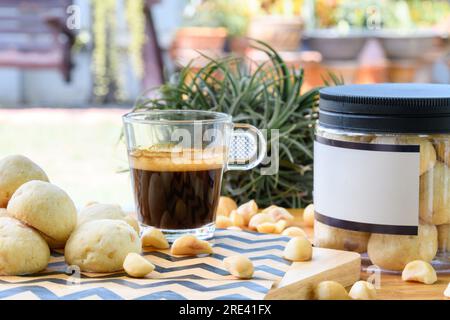 Macadamianüsse und Kekse in einer Plastikbox neben einer Tasse schwarzen Kaffees auf einem Holztisch im Garten Stockfoto