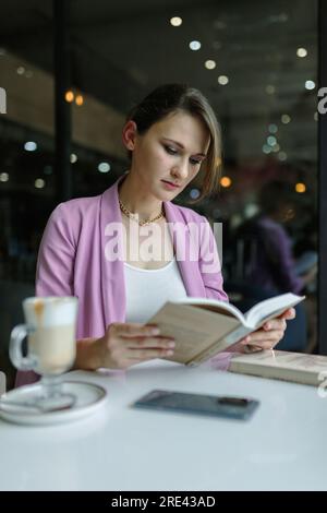 Junge, süße weiße Frau, die ein Buch in einem Café liest, Kopierraum, vertikal Stockfoto