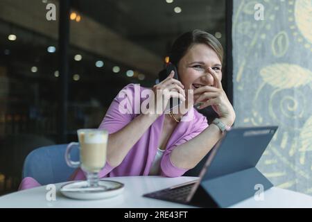 Junge weiße Geschäftsfrau, die lacht, während sie während des Mittagessens in einem Café an einem Tisch am Telefon sitzt Stockfoto