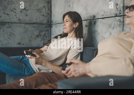 Häusliche, lustige Dame mit ihrer leitenden Mutter, die bequem auf der Couch sitzt und Joysticks hält, die Videospiele spielen. Verbringe das Wochenende zusammen. Für Spieldesign Stockfoto