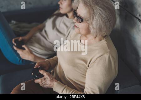 Seniorin sitzt auf dem Sofa und spielt Videospiele mit ihrer erwachsenen Tochter, die gemeinsam Freizeit verbringt, und entspannt sich am Wochenende zu Hause. Amüsiert euch Stockfoto