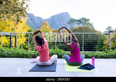 Rücken von afroamerikanischer Mutter und Tochter, die zu Hause Yoga auf der sonnigen Terrasse praktizieren Stockfoto