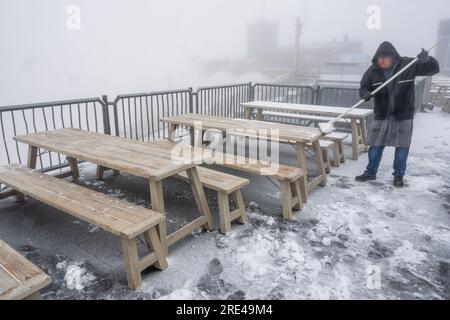 Garmisch Partenkirchen, Deutschland. 25. Juli 2023. Während eines Schneesturms auf der Zugspitze (2962 Meter) entfernt ein Mann Schnee von den Tischen auf der schneebedeckten Aussichtsplattform. Kredit: Peter Kneffel/dpa/Alamy Live News Stockfoto