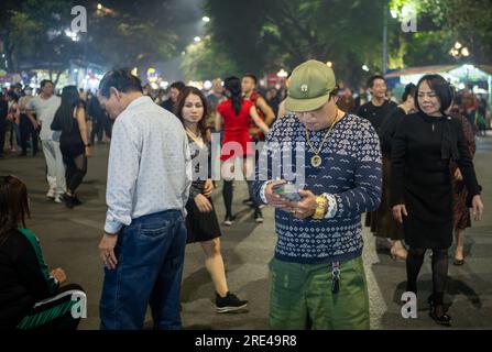 Ein vietnamesischer Mann, der schweren Gold- und Diamantschmuck, Kampfhosen und einen olivgrünen Hut trägt, überprüft sein Handy, in dem ein Fall für m steht Stockfoto