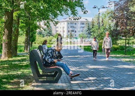 Kasan, Russland - 9. Juni 2023: Sommernachmittag im öffentlichen Park der Stadt. Mädchen mit kleinem Hund, das im Schatten auf einer Bank sitzt und auf den Bildschirm des Mobiltelefons schaut Stockfoto