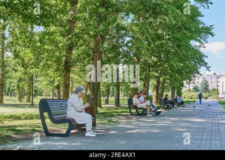 Kasan, Russland - 9. Juni 2023: Sommernachmittag im öffentlichen Stadtpark. Asiatische Seniorin, die im Schatten auf einer Bank sitzt und ein Handy benutzt. Lifestyle, te Stockfoto