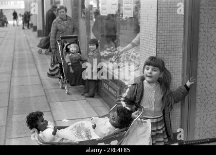 Ein junges weißes Mädchen spielt mit zwei schwarzen Kinderpuppen in einem Kinderwagen Glasgow. Schottland UK. Sie wartet vor einem Laden, während ihre Mutter drinnen einkauft. 1979 HOMER SYKES Stockfoto