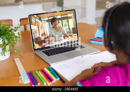 Rückansicht eines birassischen Mädchens, das Kopfhörer trägt und ein Videogespräch mit einem asiatischen Mädchen auf einem Laptop zu Hause führt Stockfoto
