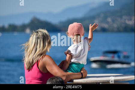 Eine Mutter und ihr Kind winkten einem Boot auf einem Bergsee zu. Eine blonde Mutter und ihre Tochter, die kleine Klamotten tragen. Stockfoto