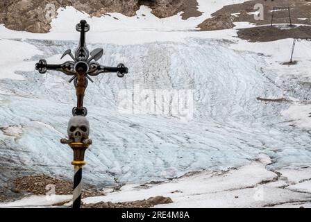 Garmisch Partenkirchen, Deutschland. 25. Juli 2023. Während einer Prozession steht ein Kreuz mit Schädel vor den Überresten des Gletschers (im Hintergrund) auf dem Zugspitz-Gletscher. Neben Informationen und Gebeten wird der Gletscher gesegnet und ein für den Anlass zusammengestelltes Bergquiem wird mit drei Sängerinnen und Windchor Premiere erhalten. Kredit: Peter Kneffel/dpa/Alamy Live News Stockfoto