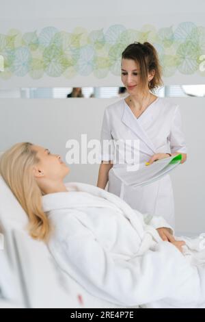 Vertikales Porträt einer Arztin, die eine Krankengeschichtsliste während der Station ausfüllt, eine junge Patientin, die vor dem Eingriff auf der Liege liegt Stockfoto