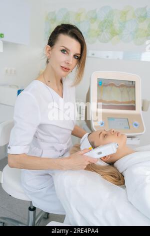 Vertikalporträt einer Kosmetikerin, die eine Ultraschall-Gesichtsliftmassage mit professioneller Ausrüstung für junge Frauen in der ästhetischen Medizin durchführt Stockfoto