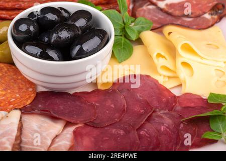 Nahaufnahme von Oliven in einer weißen Schüssel, geschnittenem Prosciutto und Käse, grünem Basilikum. Fleisch, Käse und verschiedene Wurstsorten. Stockfoto