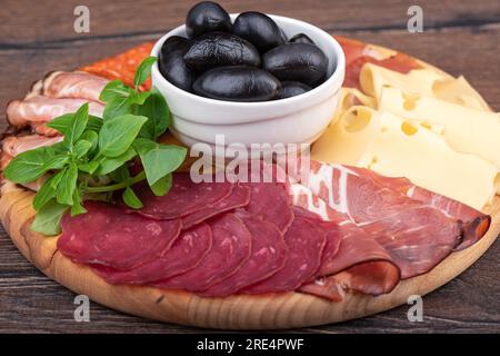 Oliven, grünes Basilikum und Teilstücke verschiedener Snacks auf einem runden Brett. Fleisch, Käse und verschiedene Wurstsorten. Schließen. Stockfoto