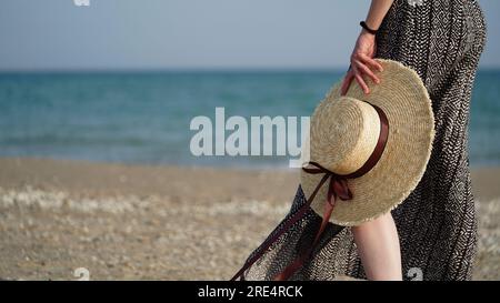 Nahaufnahme der Beine einer jungen Frau und Strohhut in der Hand auf dem Hintergrund des Sandstrandes, des Meeres, des Ozeans, des Kopierens von space.Vacation, Urlaub, Sommerurlaub concept.SE Stockfoto