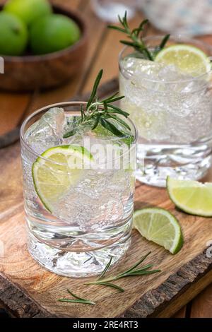 Gin- und Tonic-Cocktail garniert mit Limettenkeilen und Rosmarin auf rustikalem Holztablett Stockfoto
