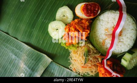 Schmackhafter Reis, Nudeln, Gurke, gewürzte Kartoffeln auf Bananenblatt. Traditionelle indonesische Speisen mit rotem weißem Band für die Feierlichkeiten zum Unabhängigkeitstag Stockfoto