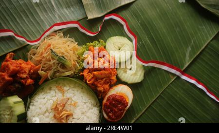 Schmackhafter Reis, Nudeln, Gurke, gewürzte Kartoffeln auf Bananenblatt. Traditionelle indonesische Speisen mit rotem weißem Band für die Feierlichkeiten zum Unabhängigkeitstag Stockfoto