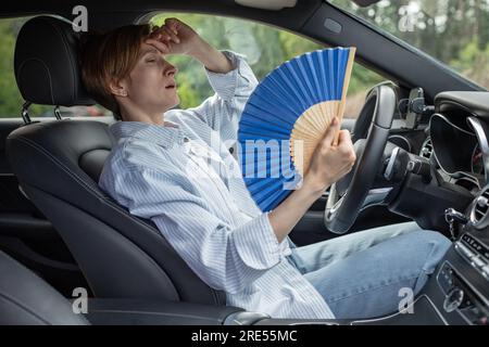 Eine müde, erschöpfte Frau mittleren Alters, die mit einem blauen Fächer wedelt, leidet an Steifigkeit im Auto. Stockfoto