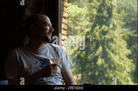 Entspannter Mann mittleren Alters mit einem Glas Weißwein in der Hand, beleuchtet durch ein gedämpftes Abendlicht mit Blick auf ein ländliches Haus mit grünem Tre Stockfoto