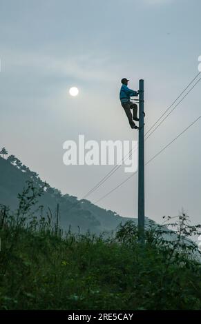 Die Silhouette eines Arbeiters, der auf einen Stab klettert, um hängende Drähte zu installieren, während die Sonne kaum durch den Smog und die Wolken in China zu sehen ist Stockfoto