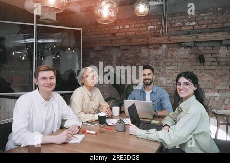 Das glückliche Business-Team sitzt im Büro und lächelt, während es in die Kamera schaut. Kollegen, die an einem Projekt arbeiten und Laptops verwenden. Der Start Stockfoto