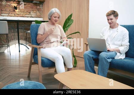 Eine Erwachsene Frau mit einem Handy und ein junger Mann mit einem Laptop, der während der Arbeit auf einem Sessel im Büro sitzt. Start, Zusammenarbeit, Management Stockfoto